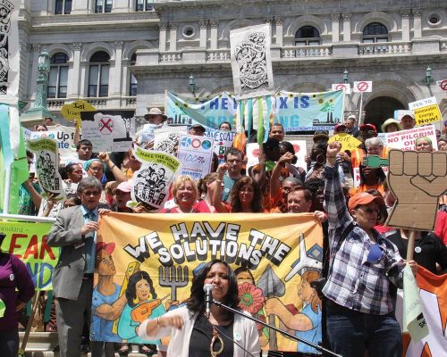 Woman speaking at climate rally with handmade signs, banner behind her says We have the solutions