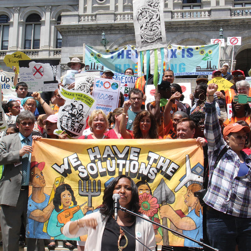 NY Renews rallying for the strongest climate bill in the country — people with signs and banners, woman speaking in front of banner that says We have the solutions