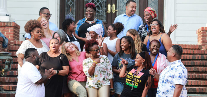 A predominately Black group of people laughing and enjoying themselves outside in front of a brick porch