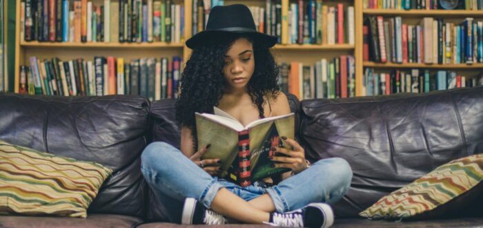 Black girl in Black brim hat, sitting on couch reading a book, in front of a large bookcase