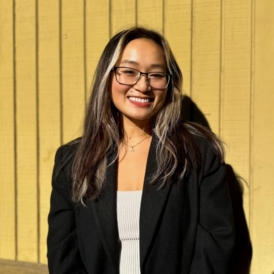 Asian woman in Black blazer and white top with red lipstick standing in front of yellow wall