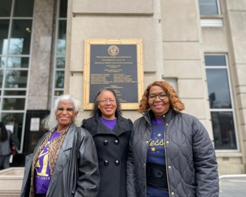 3 older Black women all dressed in black coats, standing outside side by side smiling in front of a building