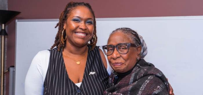 two Black women standing side by side one seemingly younger and taller wearing a pinstripe vest and smiling the other shorter and older wearing glasses and a smile