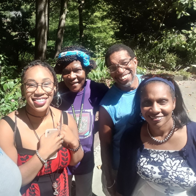 three Black women, one with glasses, and one Black man with glasses, outside in nature all with big smiles on their faces